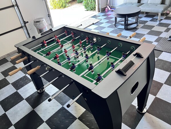 Ein Tischkicker in einem Raum mit Schachbrettmuster, einer Stehlampe und einem Roller im Hintergrund.