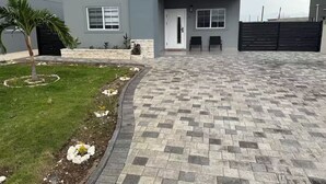 A modern house with a paved driveway, a small garden with flowers, and a palm tree.