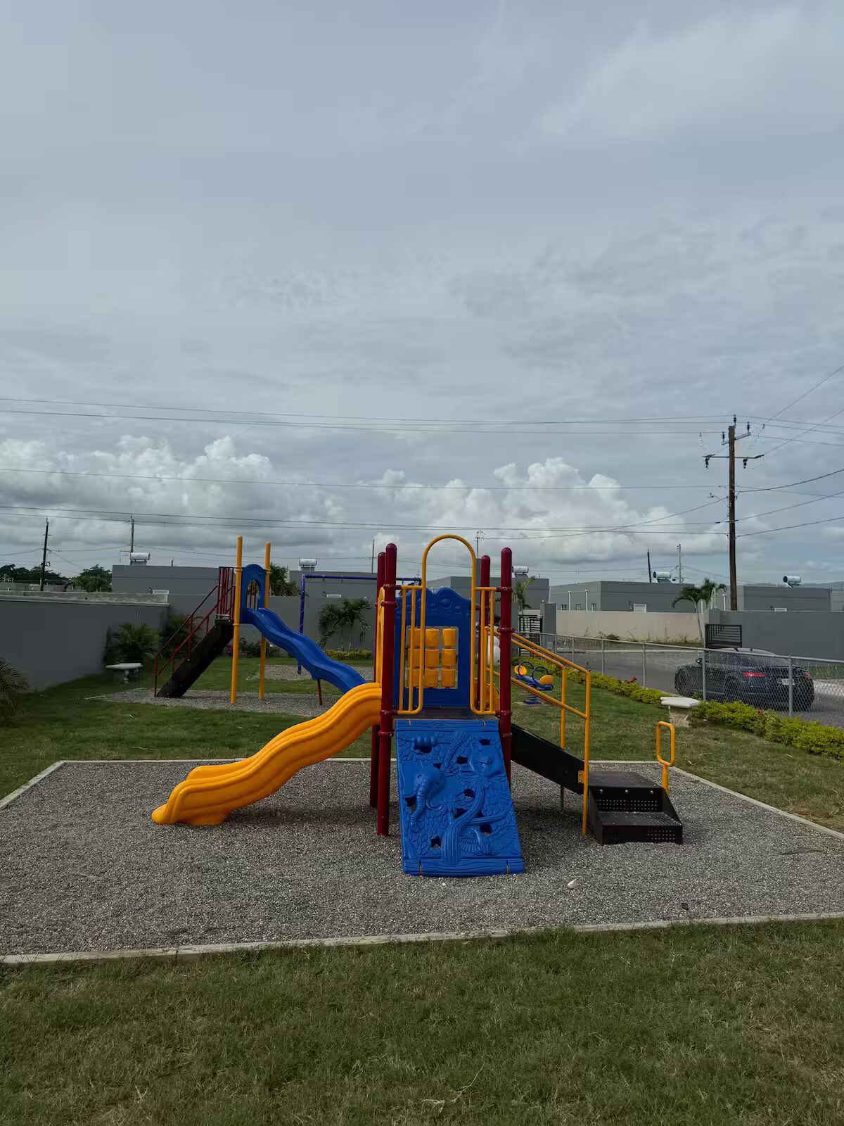 Ein Spielplatz mit Rutschen und Klettergerüsten, umgeben von Kies und Rasen, mit Gebäuden und bewölktem Himmel im Hintergrund.