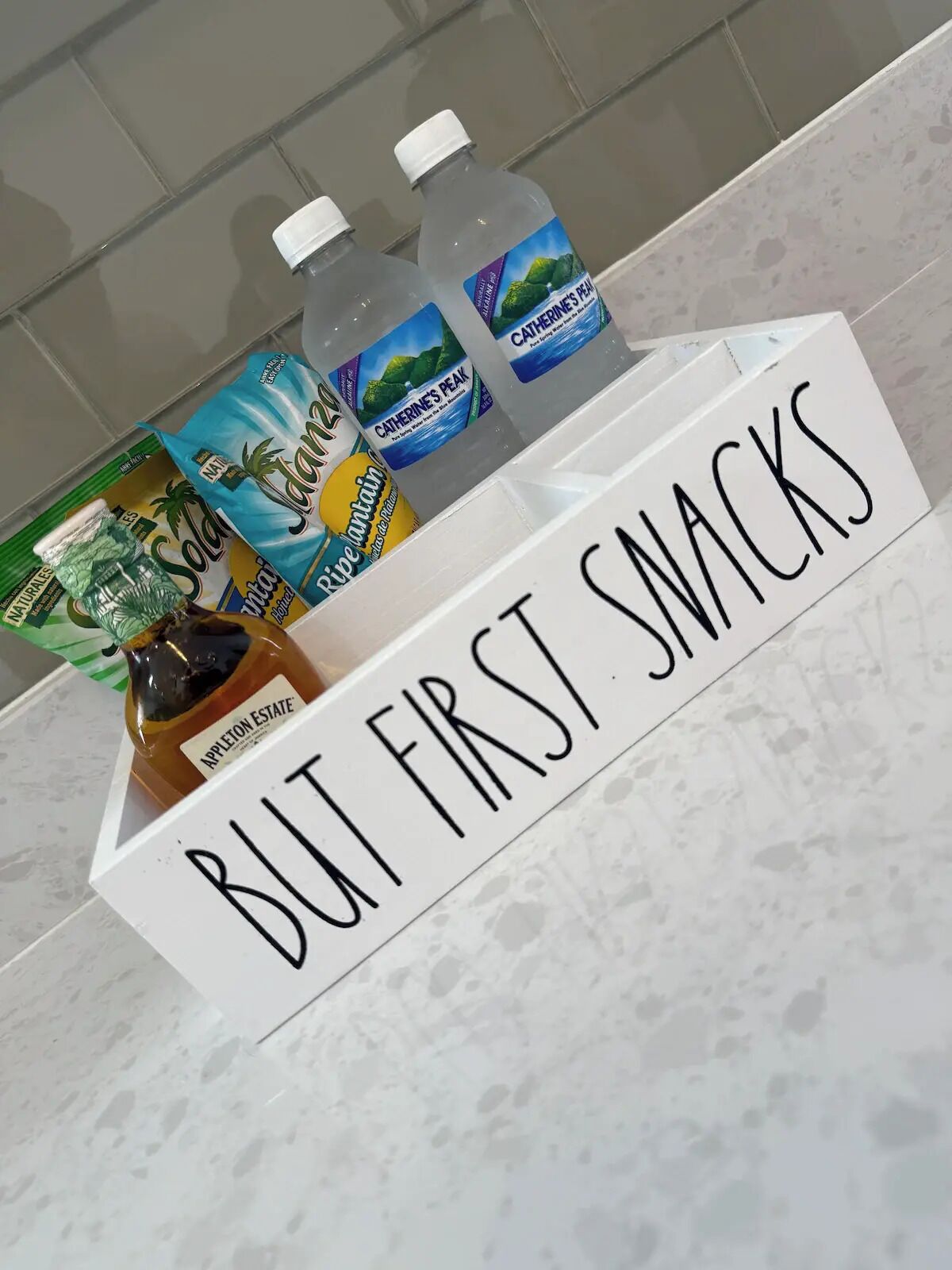 Una bandeja con un cartel que dice 'BUT FIRST SNACKS' y varias bebidas, incluyendo una botella de jugo de manzana y dos botellas de agua.