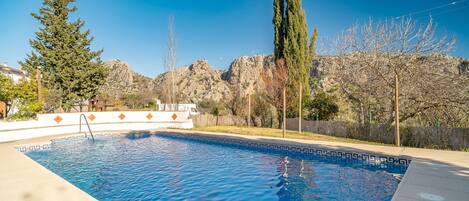 A swimming pool with clear blue water, surrounded by a concrete deck. There are trees and a rocky mountain in the background.