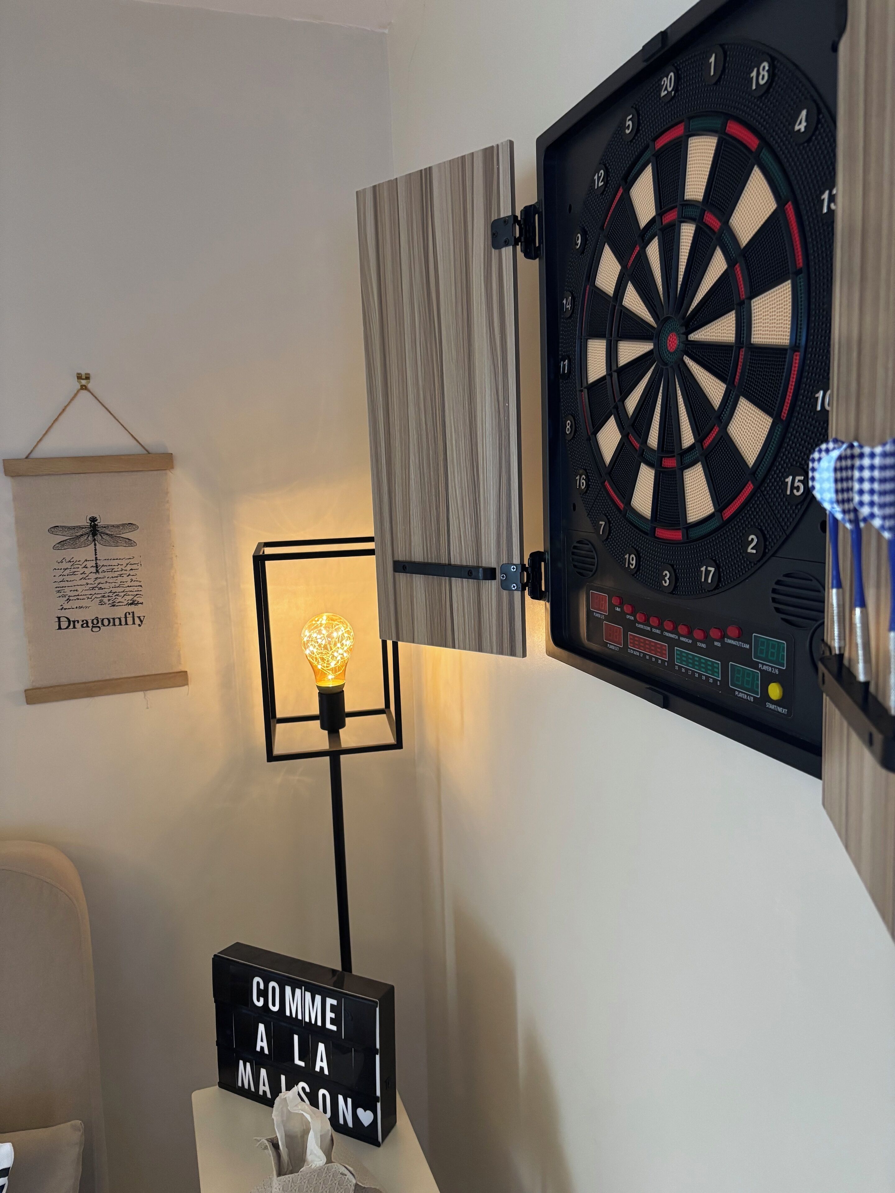 Une cible de fléchettes fixée au mur, un tableau encadré, un luminaire et un panneau avec du texte.