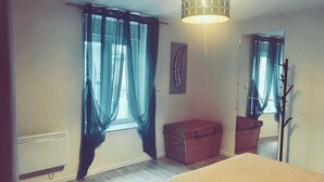 Ein Schlafzimmer mit Bett, einer Kommode, einem Korbwäscher, einem an der Wand befestigten Spiegel und transparenten Vorhängen.