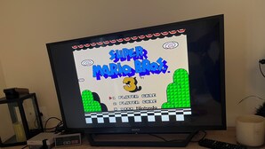 Ein Fernseher zeigt ein Videospiel, platziert auf einem Holztisch zusammen mit verschiedenen elektronischen Geräten und einer Fernbedienung.