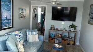Ein Wohnzimmer mit einem hellblauen Sofa, einem Holztisch, einem an der Wand montierten Fernseher und einem Deckenventilator.