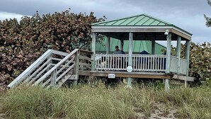 Eine Strandhütte mit einem grünen Dach und einer Holzterrasse, umgeben von hohem Gras und einem Laubhaufen.