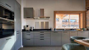 Eine moderne Küche mit Edelstahlgeräten, grauen Schränken und Blick auf eine Holzhütte durch ein großes Fenster.