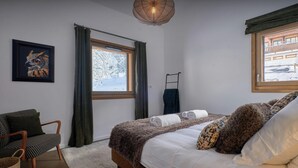 Ein Schlafzimmer mit Bett, Sessel, Fenster mit Vorhängen, einem Wandtuchhalter und einem gerahmten Bild.