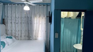 Quarto com cama, ventoinha no teto, cortinas e banheiro com pia e vaso sanitário.