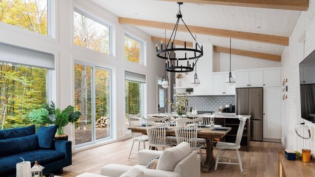 A spacious living room with a blue sofa, white armchairs, and a dining area with a wooden table and chairs. Large windows offer a view of the outdoors. - Nature & Confort: 3-bedroom cottage near Tremblant (Mont-Tremblant)