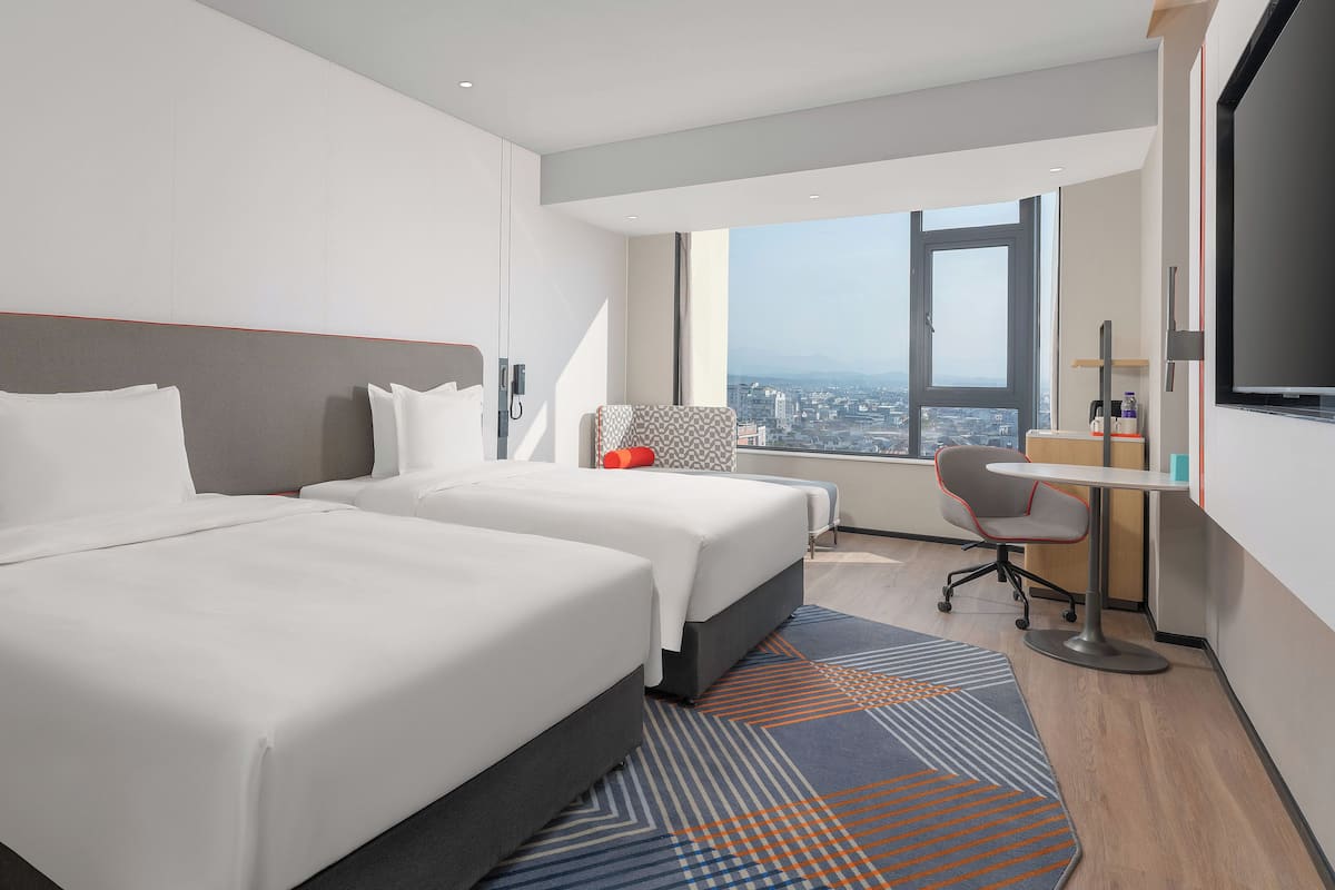 A modern hotel room with two beds, a desk, and a large window offering a city view.
