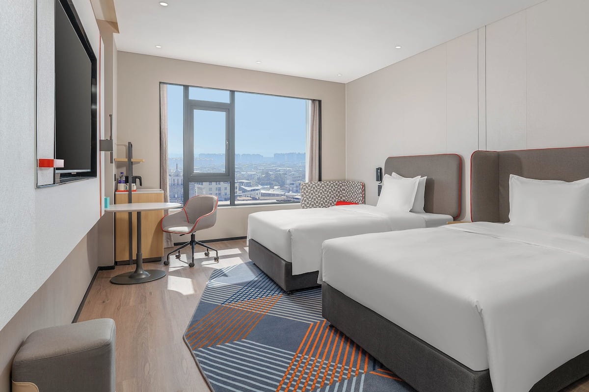 A hotel room with two beds, a desk, a chair, and a large window offering a city view.