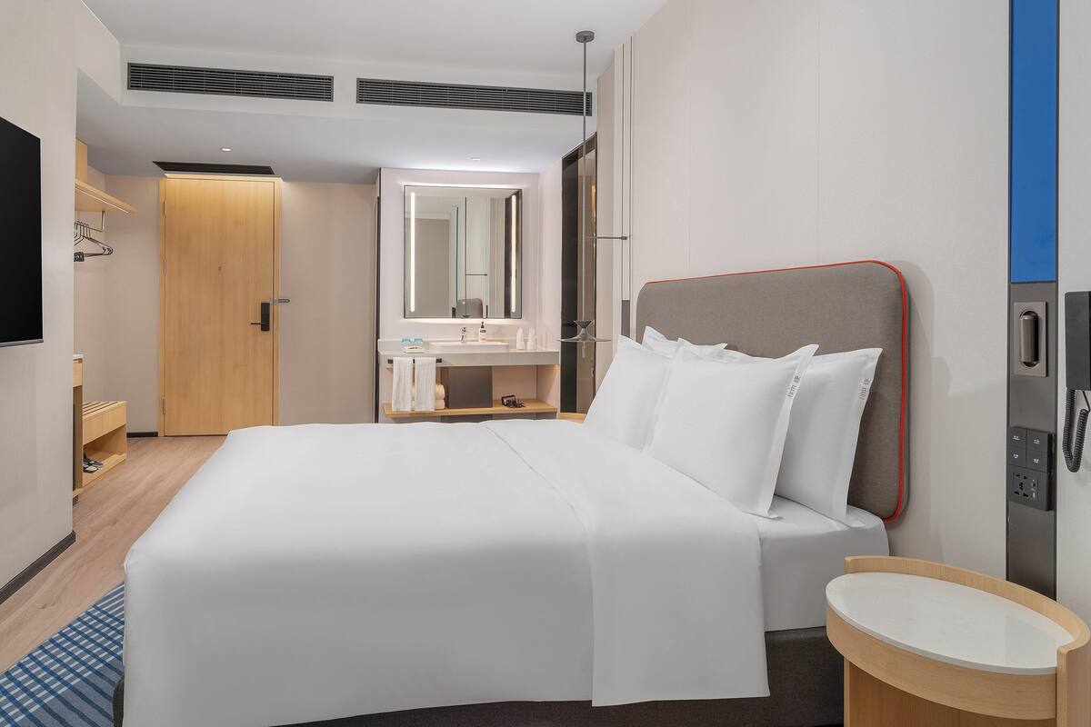 A hotel room with a bed, a bedside table, a mirror, a TV, and a bathroom with a sink and mirror.