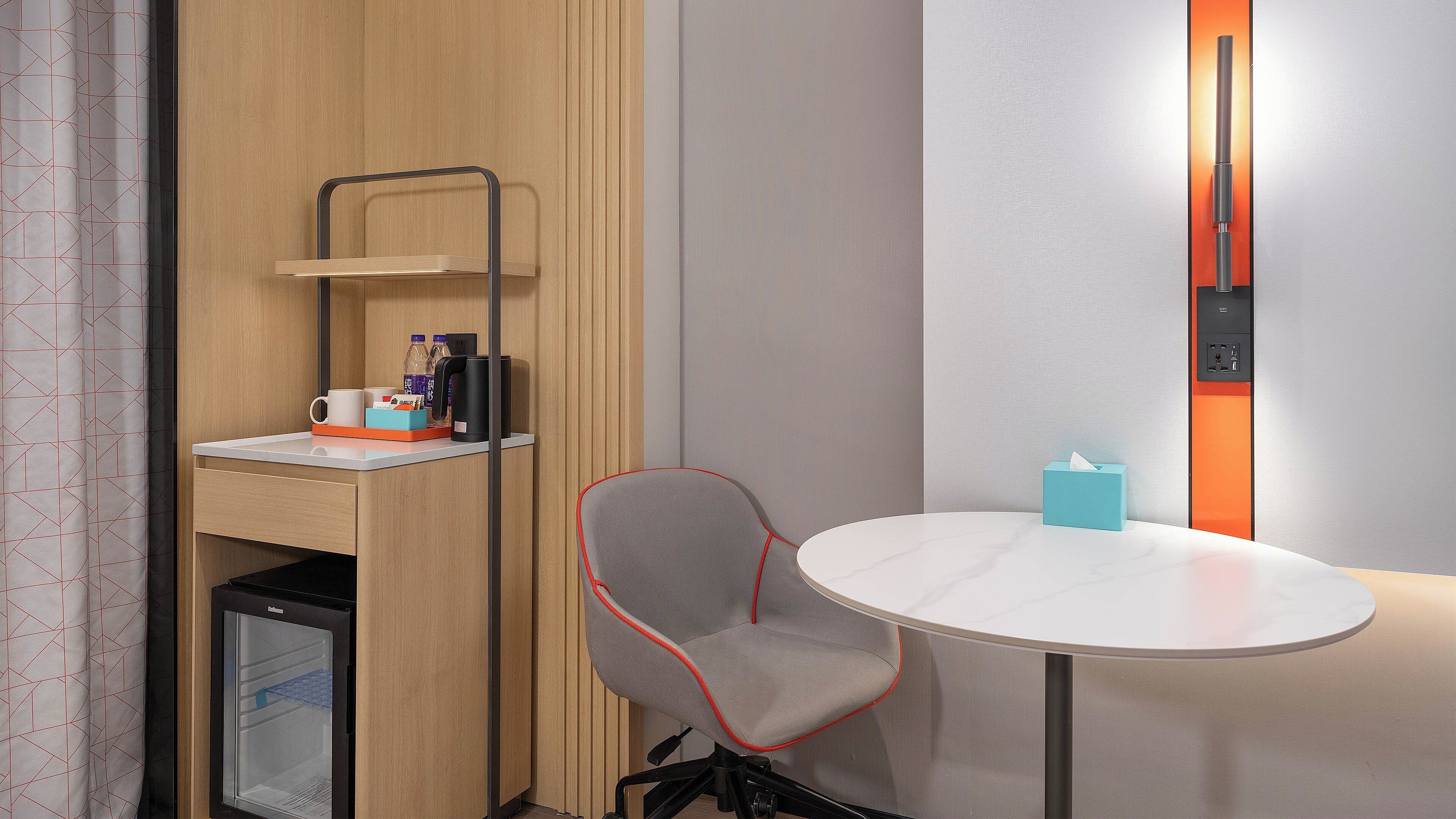 Ein kompaktes Hotelzimmer mit Küchenzeile, einem kleinen Kühlschrank, einem Sessel, einem Tisch und einem Nachttisch mit Lampe.