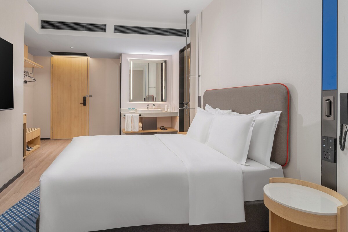 A hotel room with a bed, a bedside table, a mirror, a TV, and a bathroom with a sink and mirror.