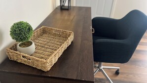 Ein dunkler Holztisch mit einer gewebten Ablage, auf der sich ein kleiner Topfpflanze und eine weiße Vase befinden. Ein schwarzer Stuhl steht rechts davon.