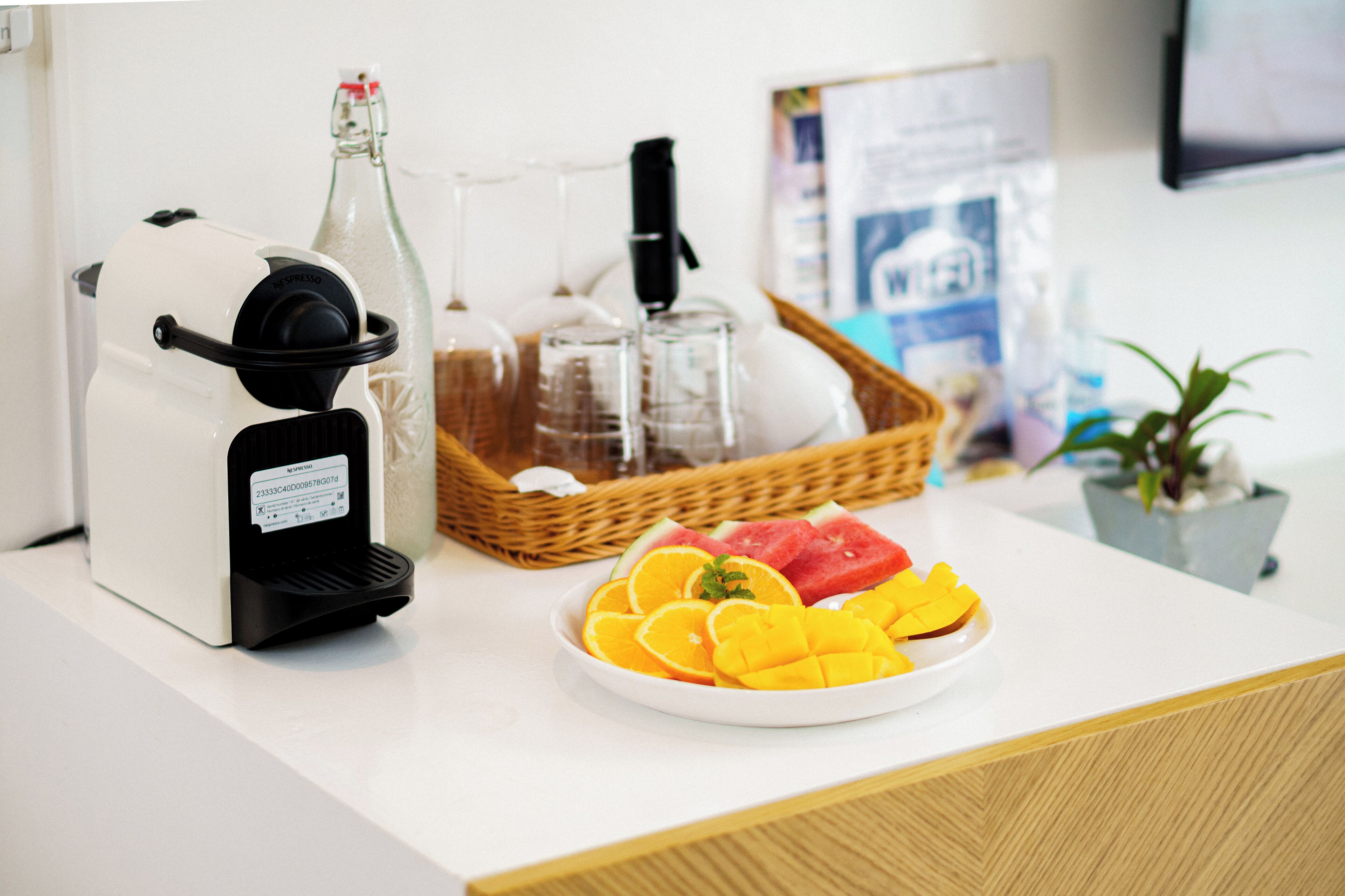 Eine Kaffeemaschine, eine Glasflasche, ein Tablett mit Gläsern und ein Teller mit Obst auf einer weißen Arbeitsplatte.