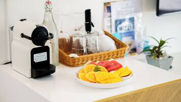 Eine Kaffeemaschine, eine Glasflasche, ein Tablett mit Gläsern und ein Teller mit Obst auf einer weißen Arbeitsplatte.