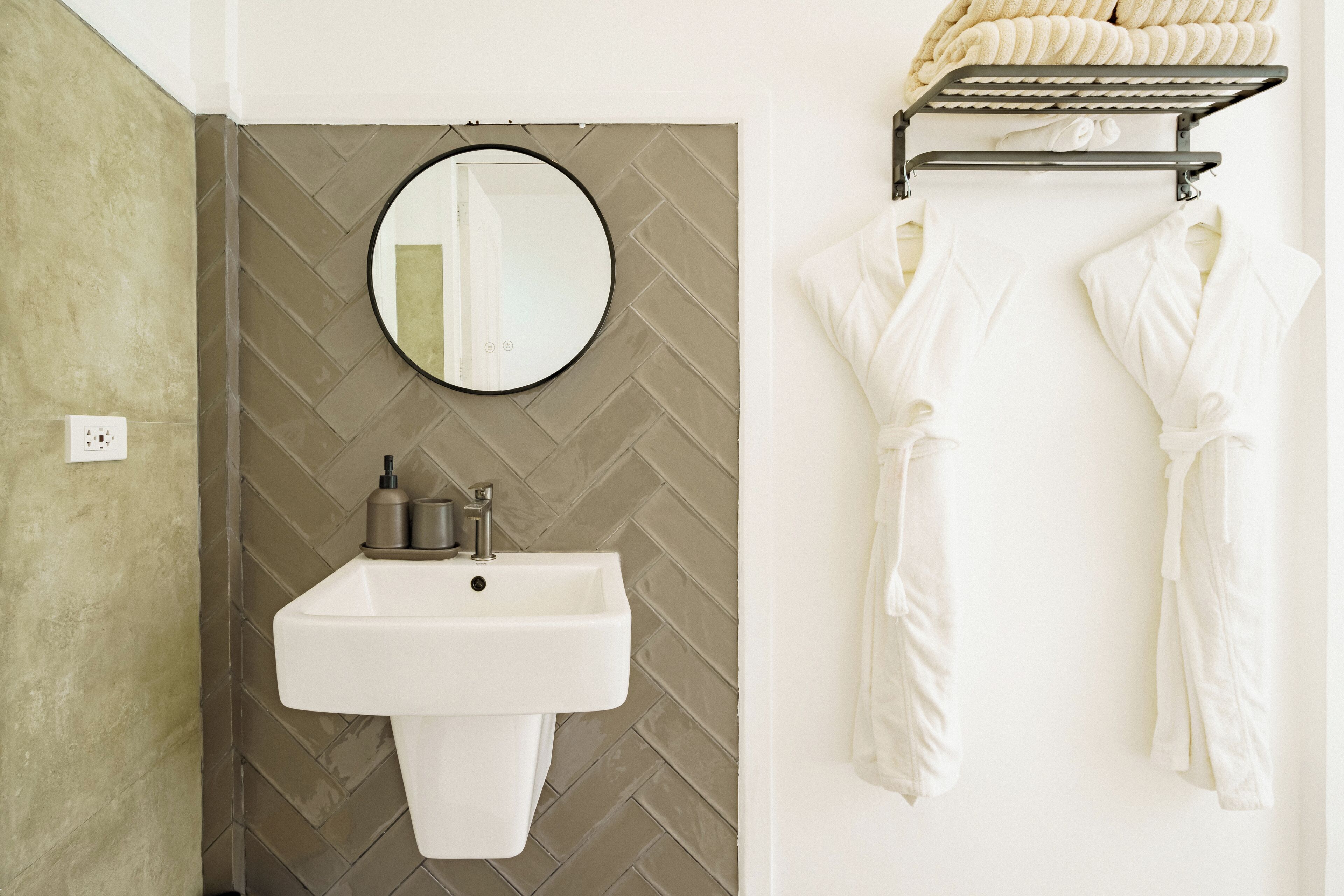 A bathroom with a white sink, a round mirror, and a towel rack with white robes.