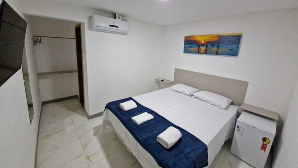 A hotel room with a bed, a small refrigerator, a painting on the wall, and an air conditioning unit.