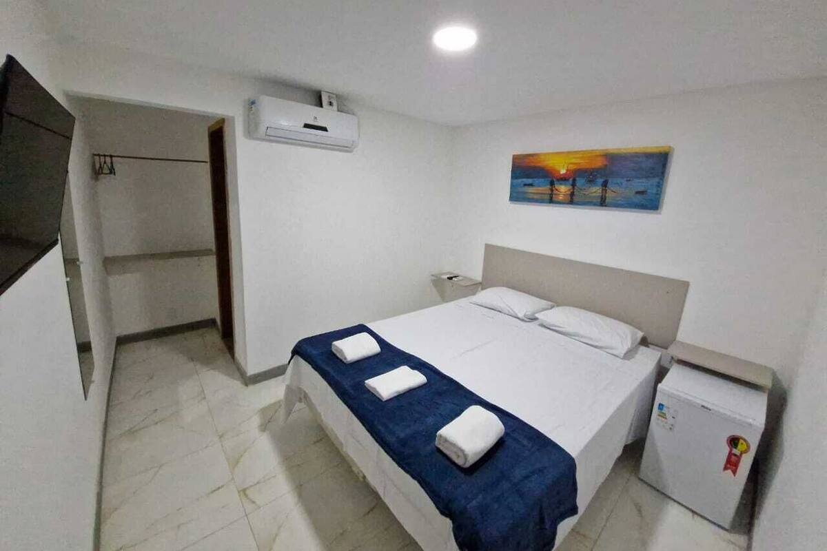 A hotel room with a bed, a small refrigerator, a painting on the wall, and an air conditioning unit.