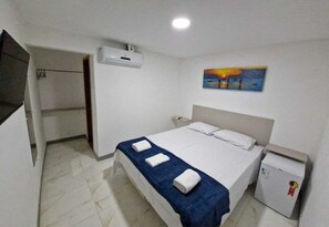 A hotel room with a bed, a small refrigerator, a painting on the wall, and an air conditioning unit. - Pousada Villa Margarida (Búzios)