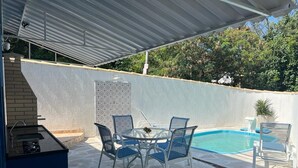 An outdoor patio with a pool, a covered grill area, and a seating arrangement.
