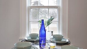 A dining table set with plates, bowls, a blue bottle, and glasses, positioned in front of a window with a view of trees.