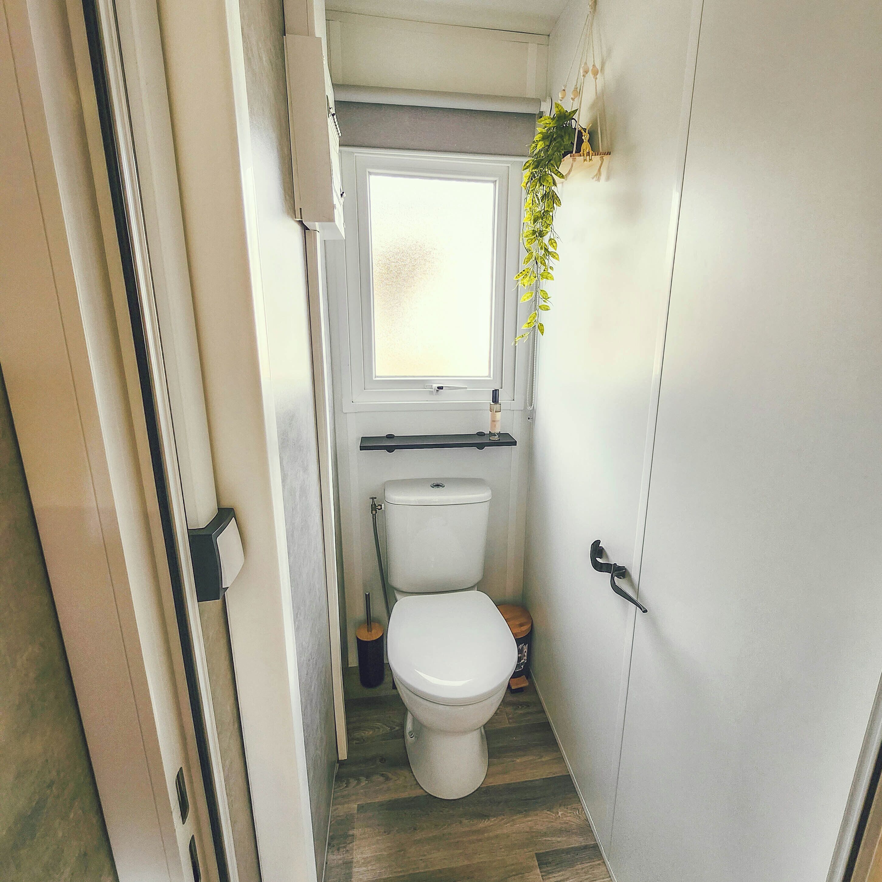 A compact bathroom with a toilet, a window, and a small shelf with a plant.