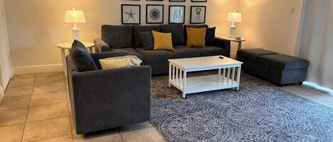 A living room with a grey sofa, a white coffee table, and a patterned rug. There are framed pictures on the wall and two lamps on side tables.