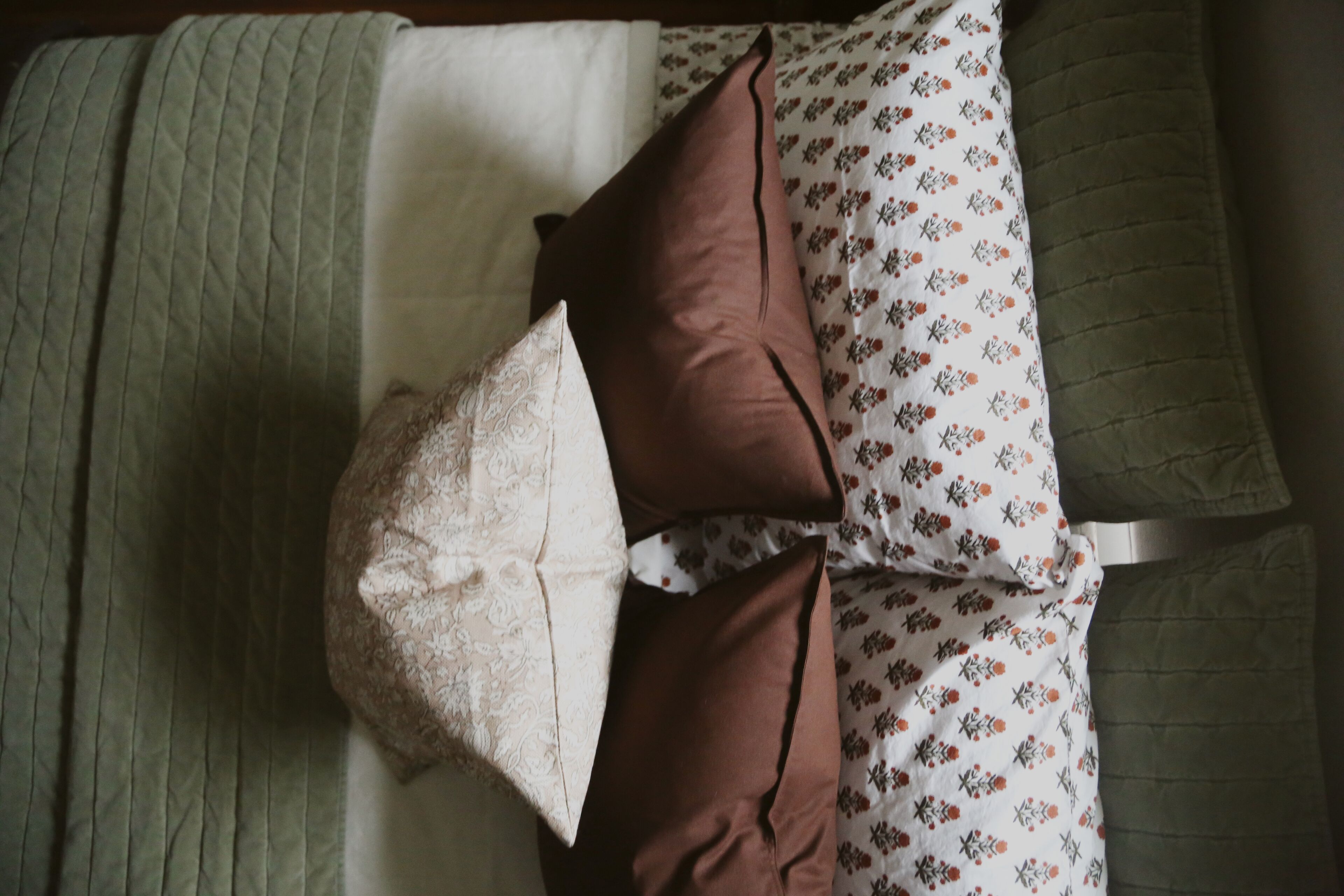 Un letto con una coperta a fantasia, una coperta verde trapuntata e due cuscini, uno marrone e uno bianco.