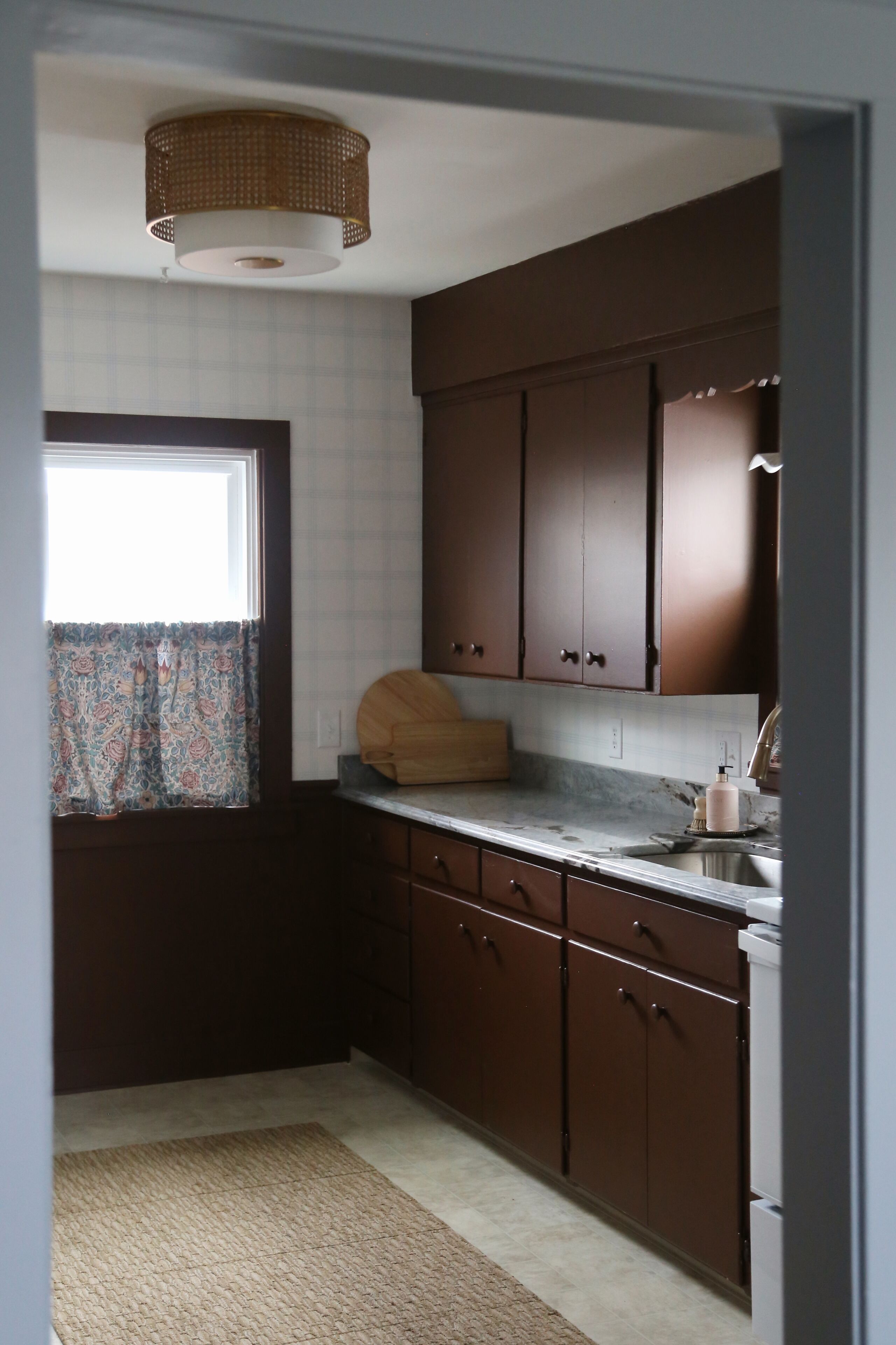 A kitchen with dark brown cabinets, a marble countertop, a window with a patterned curtain, and a woven ceiling light fixture.