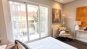 A hotel room with a large bed, a chair, a desk, and a view through a sliding door to the outside.
