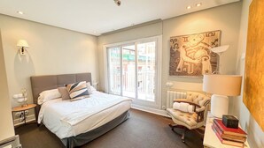 A hotel room with a large bed, a chair, a table with books, a radiator, and a painting on the wall.