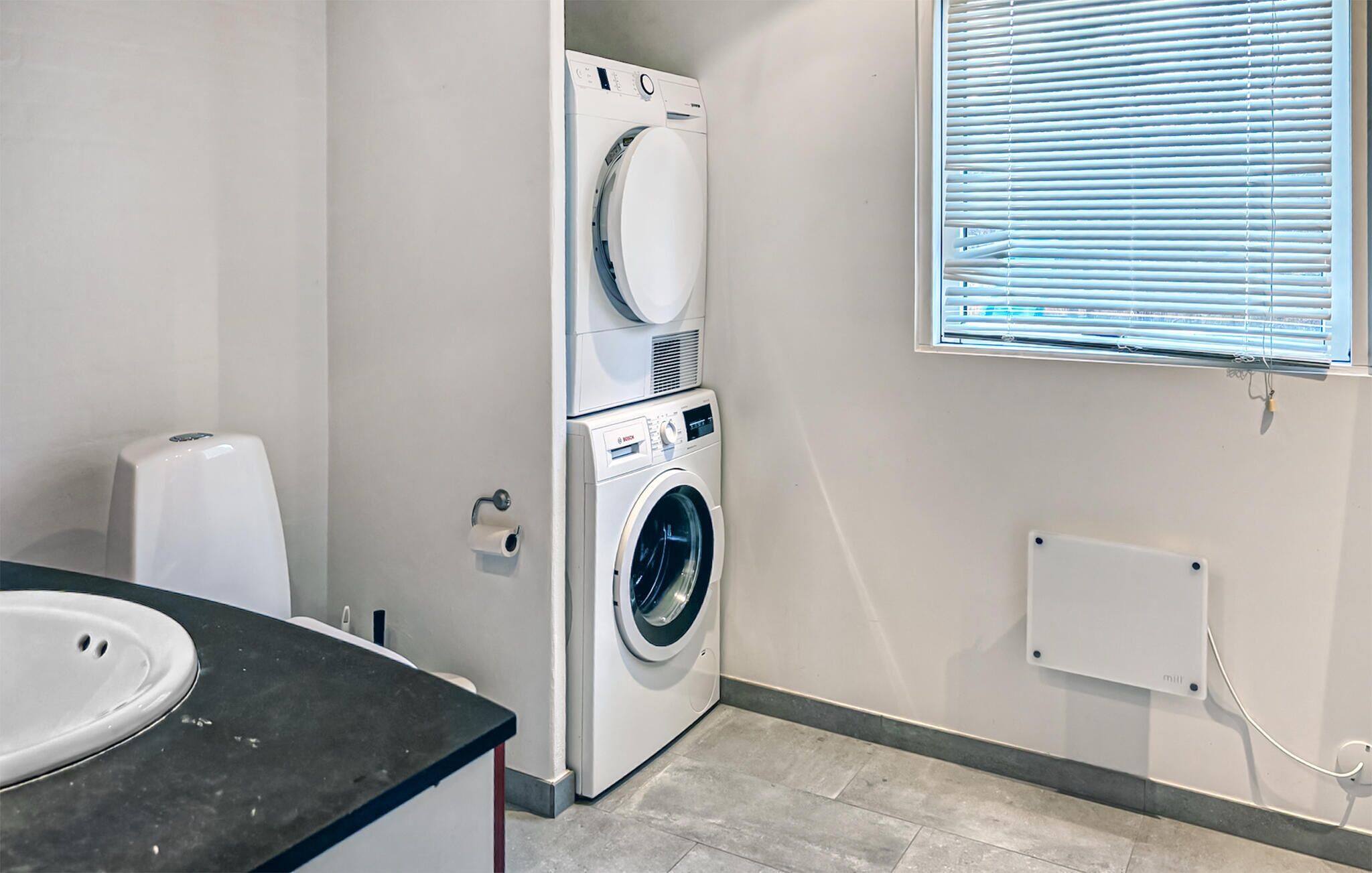 A laundry room with stacked washers and dryers, a toilet paper dispenser, and a window with blinds.