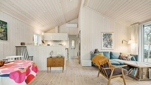 Ein geräumiger Wohnbereich mit Holzboden, einer Couch, einem Sessel und einem Tisch mit bunten Tischdecken. Im Hintergrund ist eine Küche mit weißen Schränken und einem Fenster zu sehen.