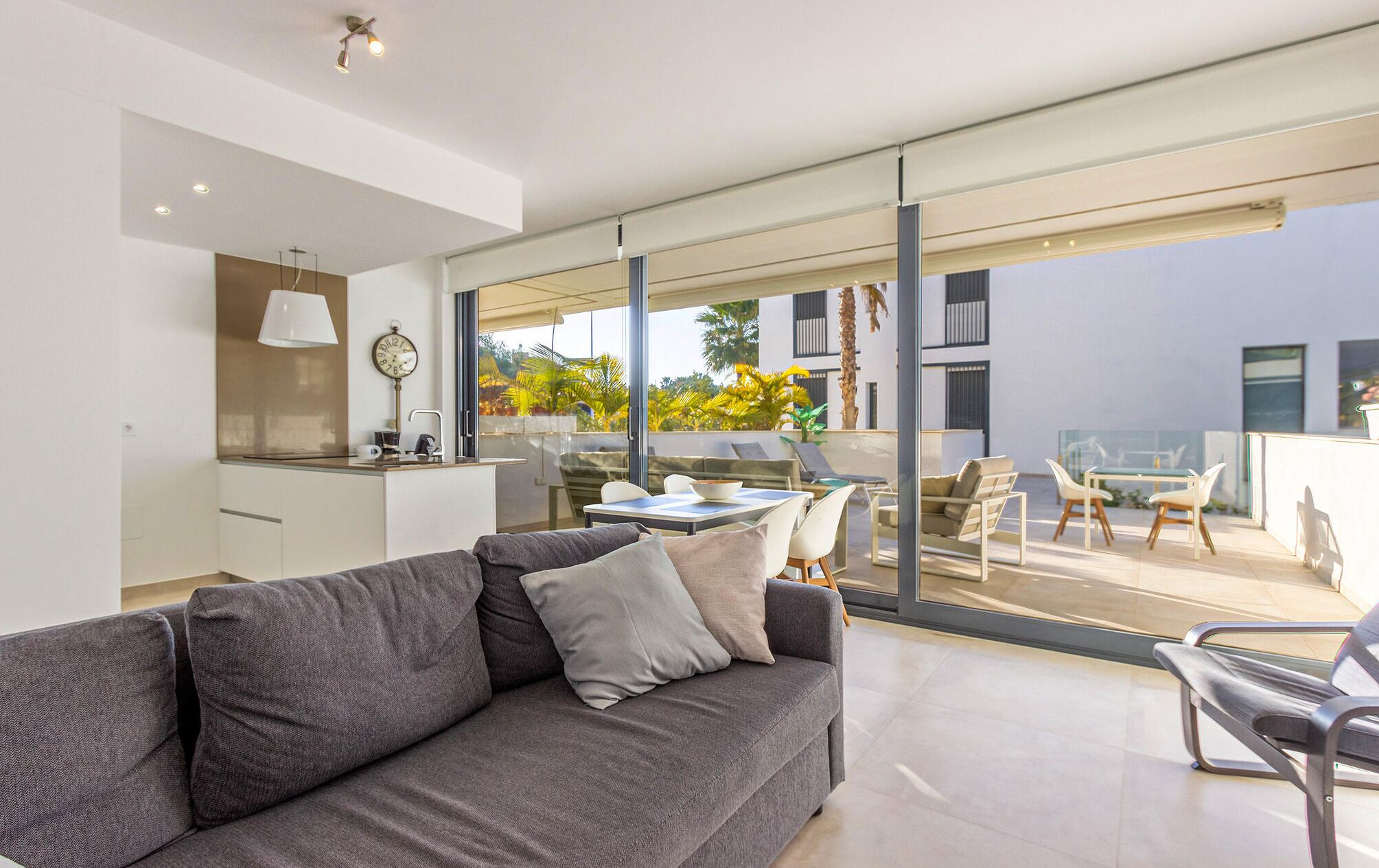 A modern living room with a grey sofa, a dining area with a table and chairs, and a large sliding glass door leading to an outdoor patio with a view of palm trees and a building.
