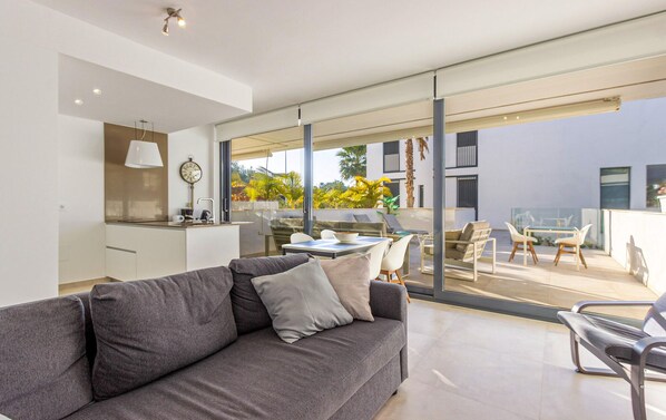 En moderne stue med en grå sofa, et spiseområde med bord og stole, samt en stor skydedør der fører ud til en udendørs terrasse med udsigt til palmer og en bygning.