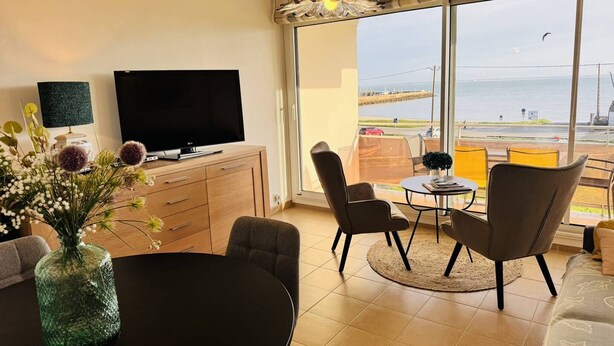 Ein modernes Wohnzimmer mit Flachbildfernseher, Esstisch und Stühlen, einem großen Fenster mit Meerblick und einem Balkon.