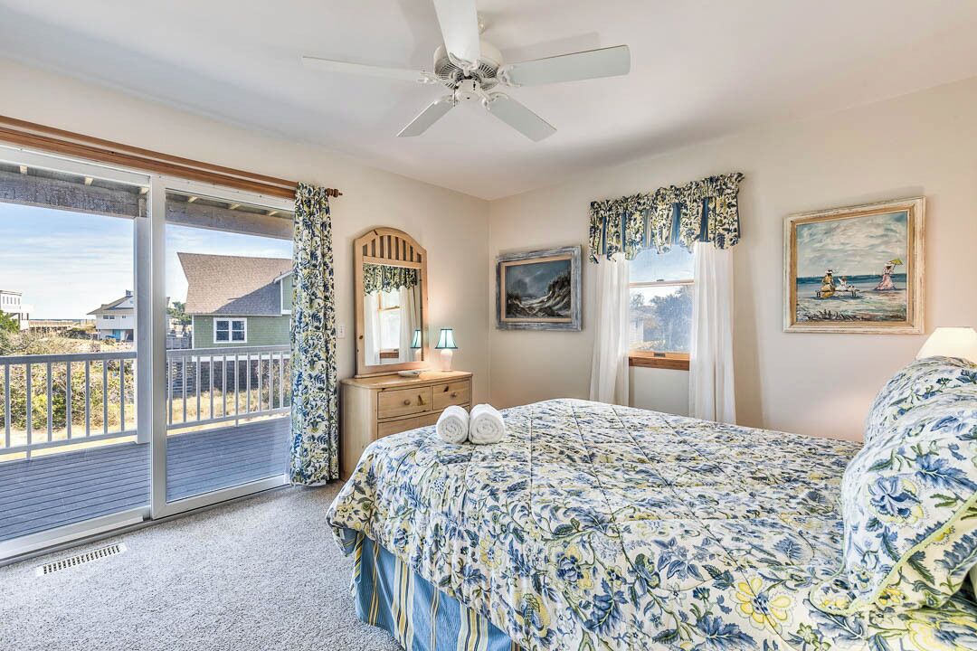 Une chambre avec un grand lit, un ventilateur de plafond, une commode, un miroir et un balcon donnant sur des maisons.