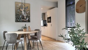 A modern dining area with a wooden table and woven chairs, a kitchen in the background, and a large window with blinds.
