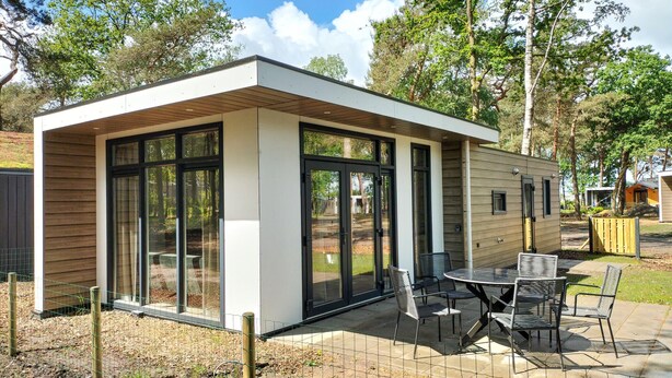 A modern, single-story mobile home with large glass doors and windows, a patio with outdoor furniture, and a natural surrounding.