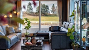 A modern living room with a sofa, armchair, and coffee table, large windows with a view of a grassy area and trees, and a glass cabinet with decorative items. - THE HORIZON RESIDENCE (Makubetsu)