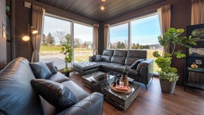 A modern living room with leather sofas, a coffee table, and large windows offering a view of the outdoors. - THE HORIZON RESIDENCE (Makubetsu)