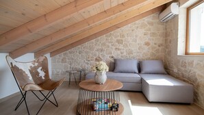 A cozy attic room with a stone wall, a wooden ceiling, a grey sofa, a wooden coffee table with books, and a chair.