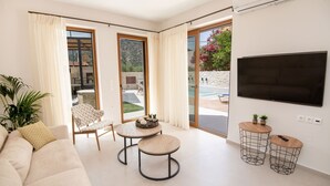 A modern living room with a sofa, two round tables, a TV, and a sliding glass door leading to an outdoor area with a pool.