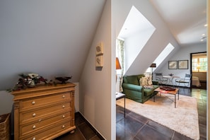 Une pièce comprenant une commode en bois, un canapé vert et une table à manger avec des chaises. Un puits de lumière se trouve au-dessus du canapé.