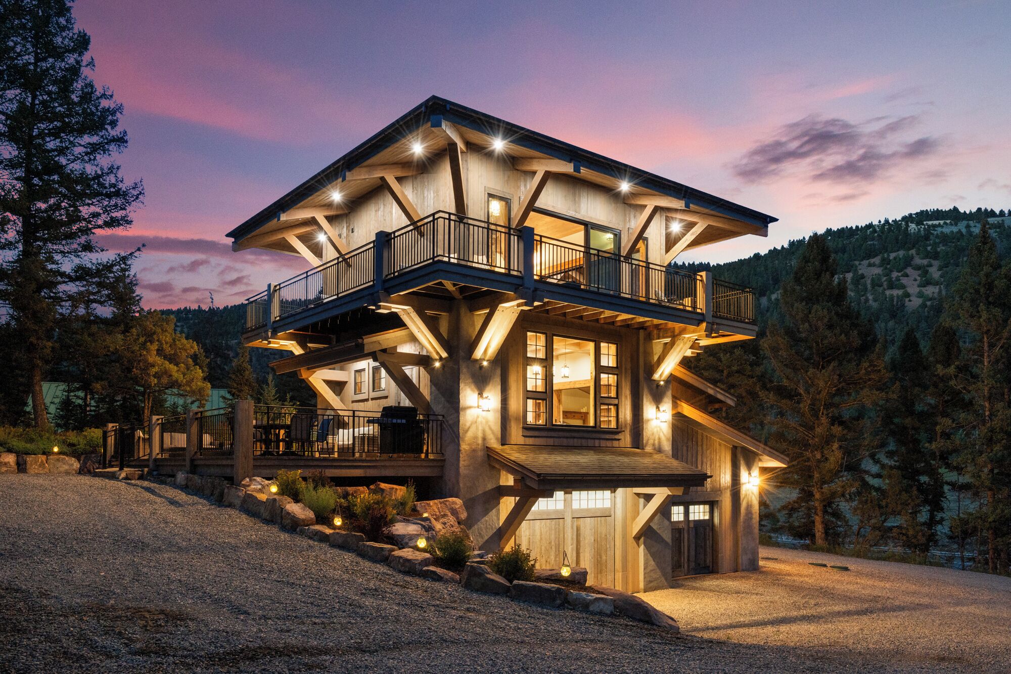 A multi-level wooden cabin with a balcony, surrounded by a gravel driveway and trees.