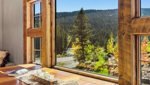 Ein Zimmer mit großen, holzrahmenverzierten Fenstern, Holzboden und Blick auf eine bewaldete Berglandschaft.