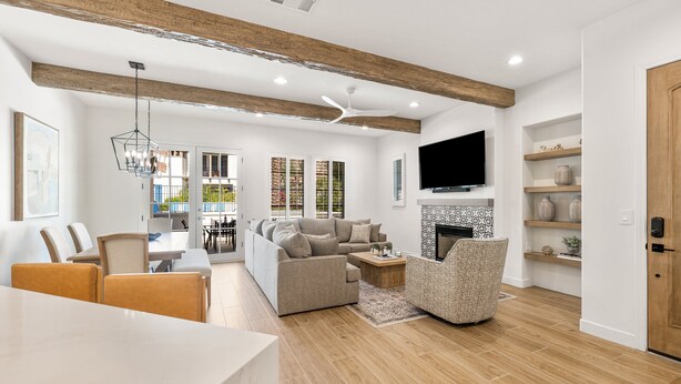 A modern living room with a sofa, coffee table, and TV. There is a dining area with chairs and a table. The room has wooden beams on the ceiling and a fireplace. - (L75) Newly Renovated Single Story Townhome with Gorgeous Views, sleeps 8 (La Quinta)